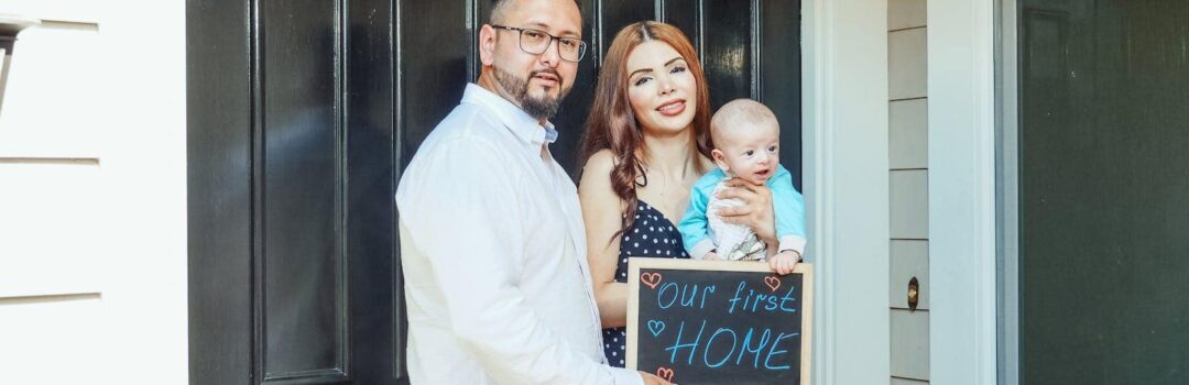 Young family with baby holding a sign 'Our First Home' at doorstep, symbolizing new beginnings.
