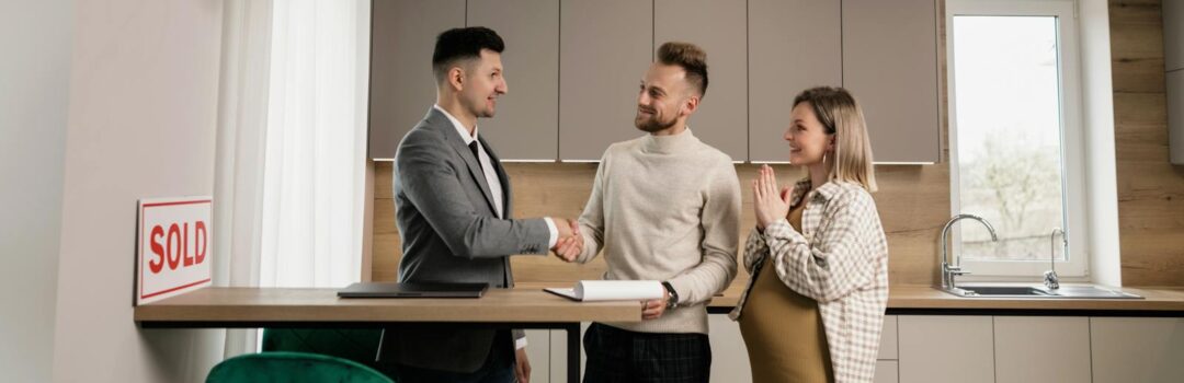A smiling couple and a realtor finalize a house purchase in a modern kitchen setting, sealing the deal with a handshake.