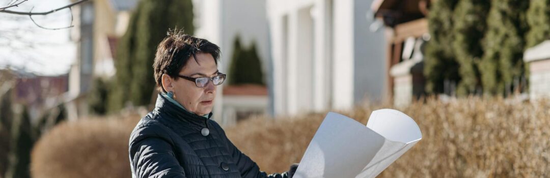 Woman examining a blueprint in a sunny residential area, considering properties.